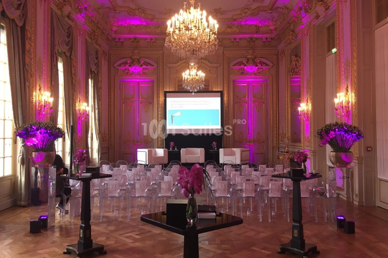 Salle de conférence élégante avec décorations florales, chaises transparentes et écran de présentation sous des lustres.