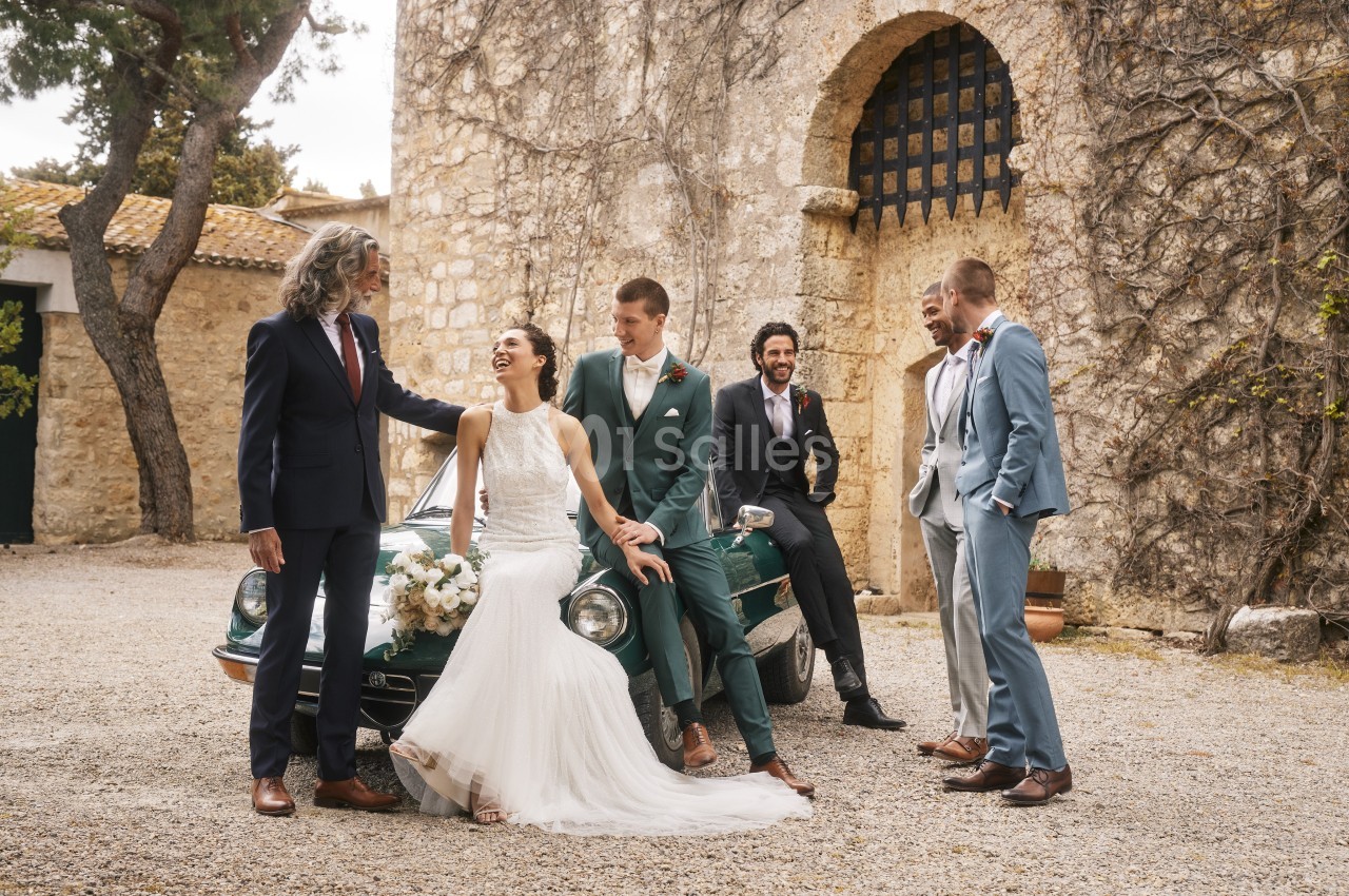 Un couple de mariés pose avec des invités élégants devant une voiture ancienne et un bâtiment en pierre.