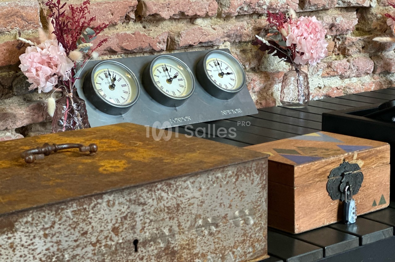 Trois boîtes en bois et métal devant un mur de briques, avec un baromètre et des fleurs en arrière-plan.
