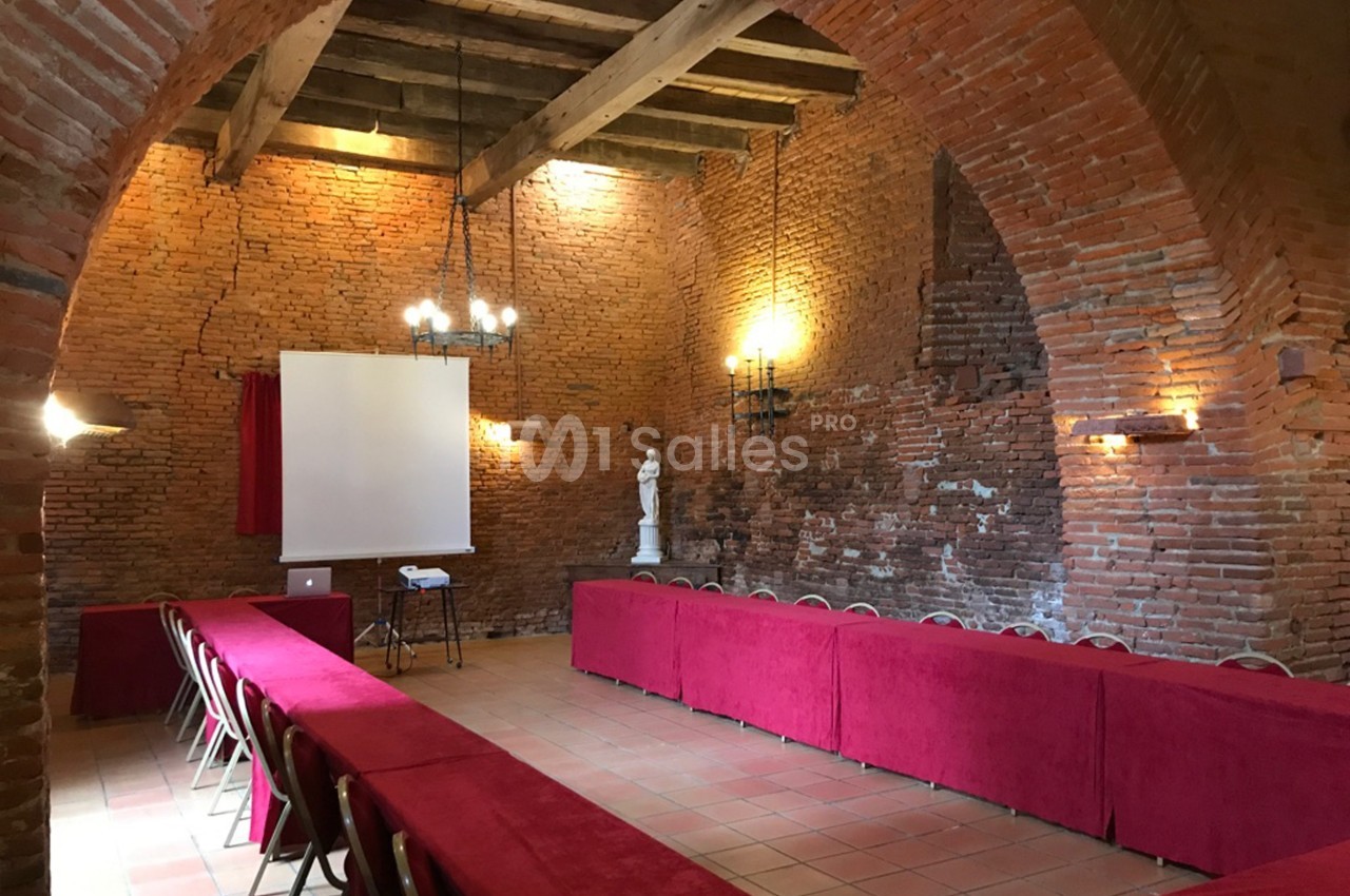 Salle en briques avec voûtes, tables recouvertes de nappes rouges, chaises alignées et écran de projection.