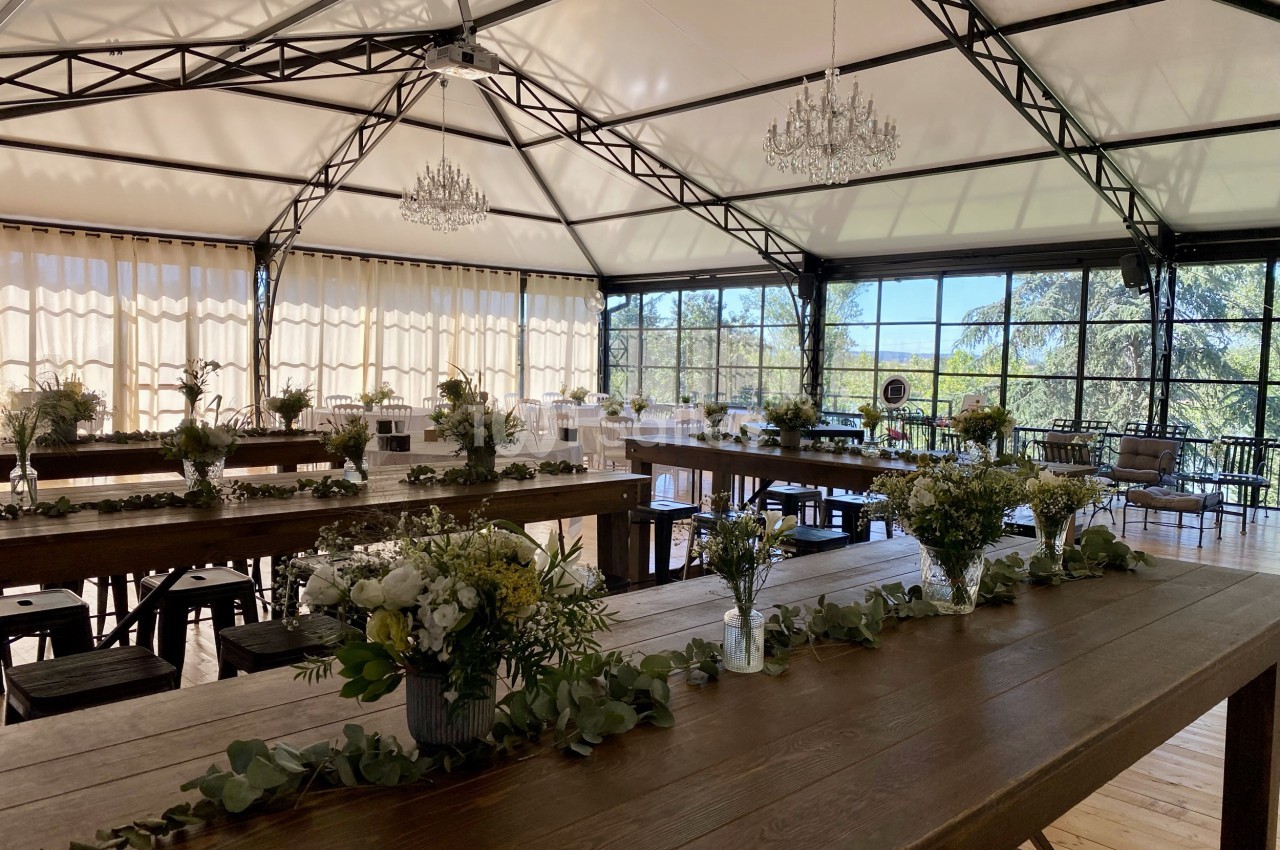 Salle lumineuse avec de grandes baies vitrées, tables en bois décorées de fleurs et guirlandes végétales.