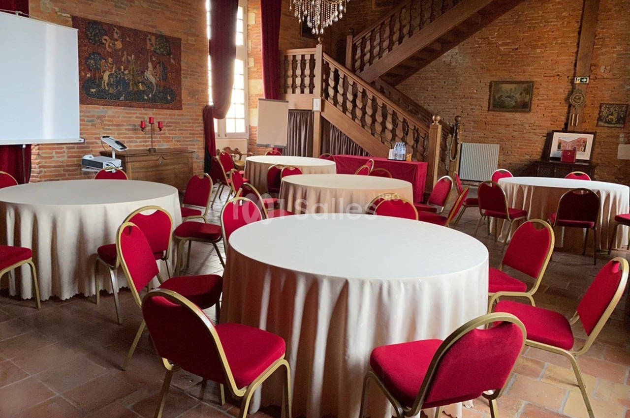 Salle avec des tables rondes nappées de blanc, entourées de chaises rouges, décorée de murs en briques et d'un lustre.