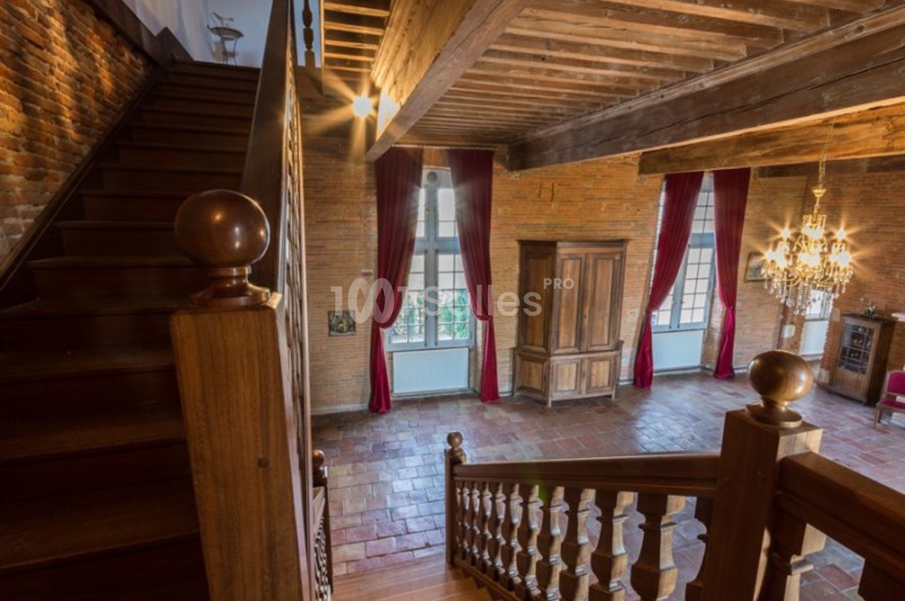 Escalier en bois menant à une pièce lumineuse avec murs en briques, grandes fenêtres, rideaux rouges et lustre.