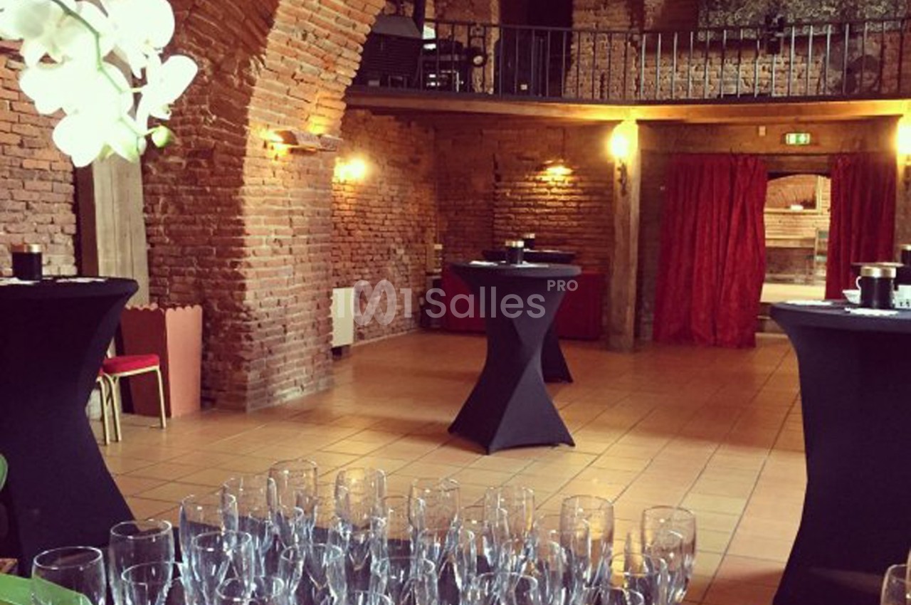 Salle en briques avec tables hautes drapées de noir, verres disposés au premier plan et mezzanine en arrière-plan.