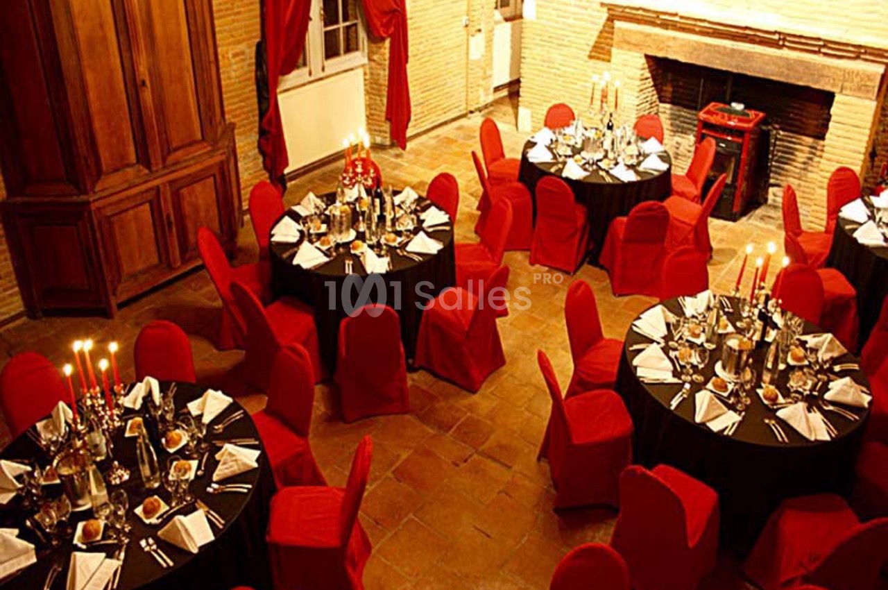 Salle de réception avec tables rondes dressées, nappes noires, chaises rouges et chandeliers allumés.