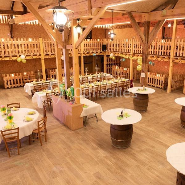 Salle de réception rustique avec tables rondes dressées, tonneaux en bois et décoration lumineuse suspendue.