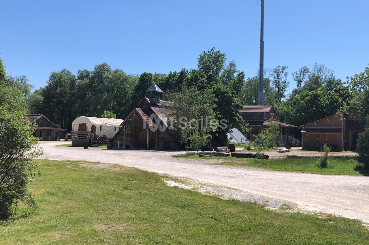 Village en plein air avec bâtiments en bois, une roulotte et une antenne, entouré de verdure sous un ciel dégagé.