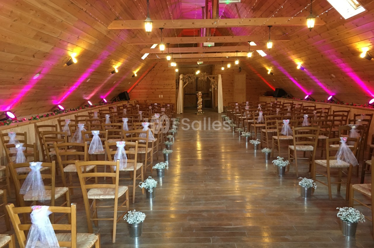 Salle en bois décorée pour une cérémonie, avec des rangées de chaises ornées de rubans blancs et des fleurs au sol.