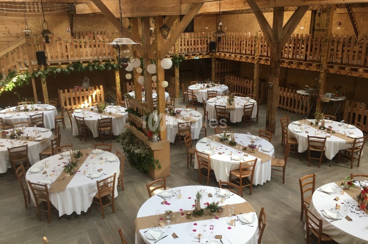 Salle de réception en bois avec tables rondes décorées de nappes blanches, chaises en bois et éléments floraux.