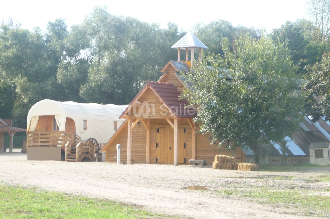 Bâtiments en bois avec une chapelle, une roulotte et des bottes de foin, entourés de verdure.