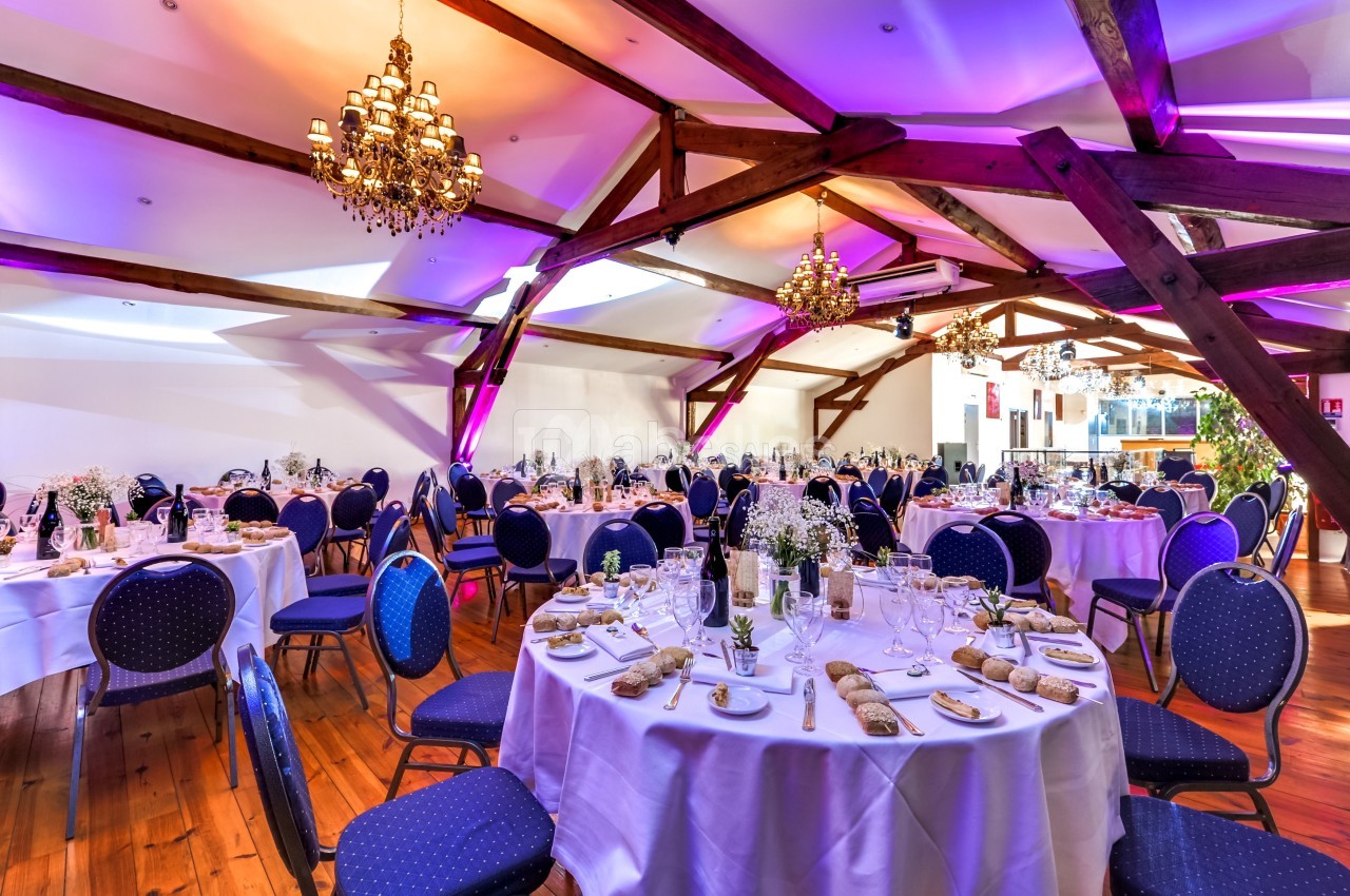 Salle de réception élégante avec tables rondes dressées, chaises bleues et éclairage chaleureux sous des poutres apparentes.