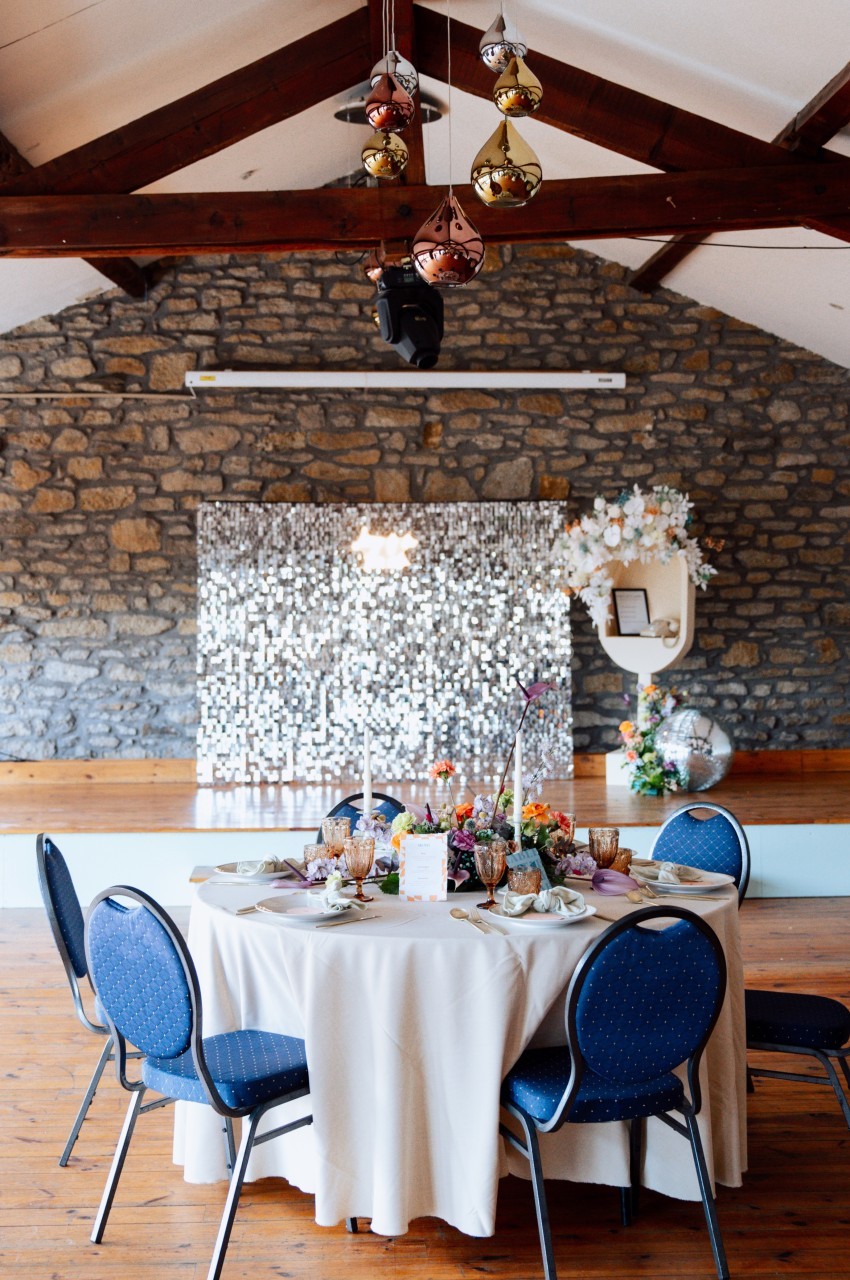 Table ronde décorée avec vaisselle colorée et fleurs, devant un mur en pierre et un fond scintillant dans une salle…