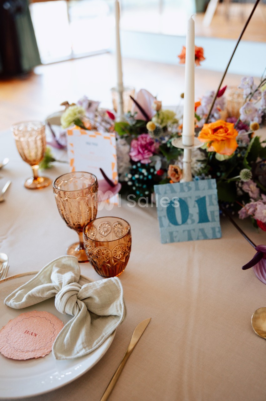 Table dressée avec vaisselle élégante, serviette nouée, verres ambrés et centre de table floral coloré.