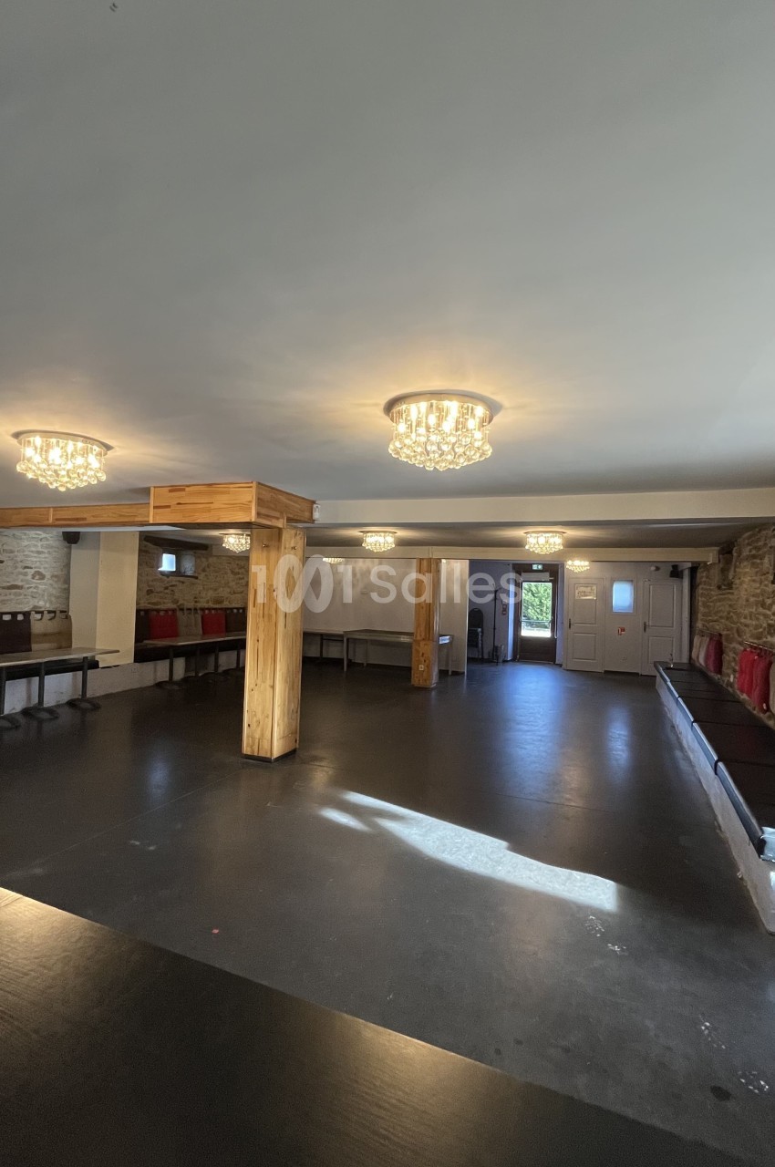 Salle vide avec sol sombre, murs en pierre, colonnes en bois, banquettes rouges et plafonniers lumineux.