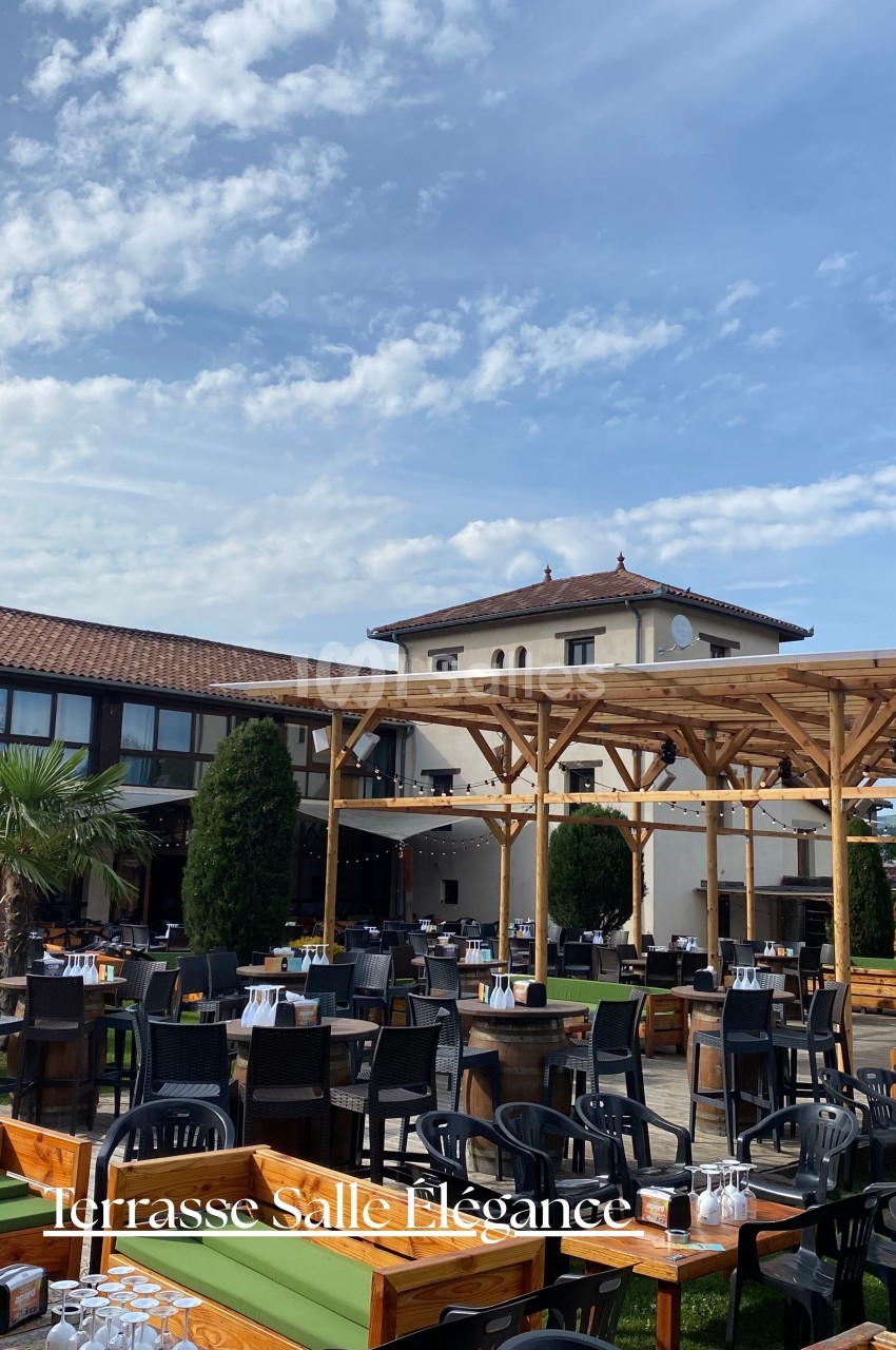 Terrasse extérieure avec tables dressées, pergola en bois et bâtiment à toit en tuiles en arrière-plan.