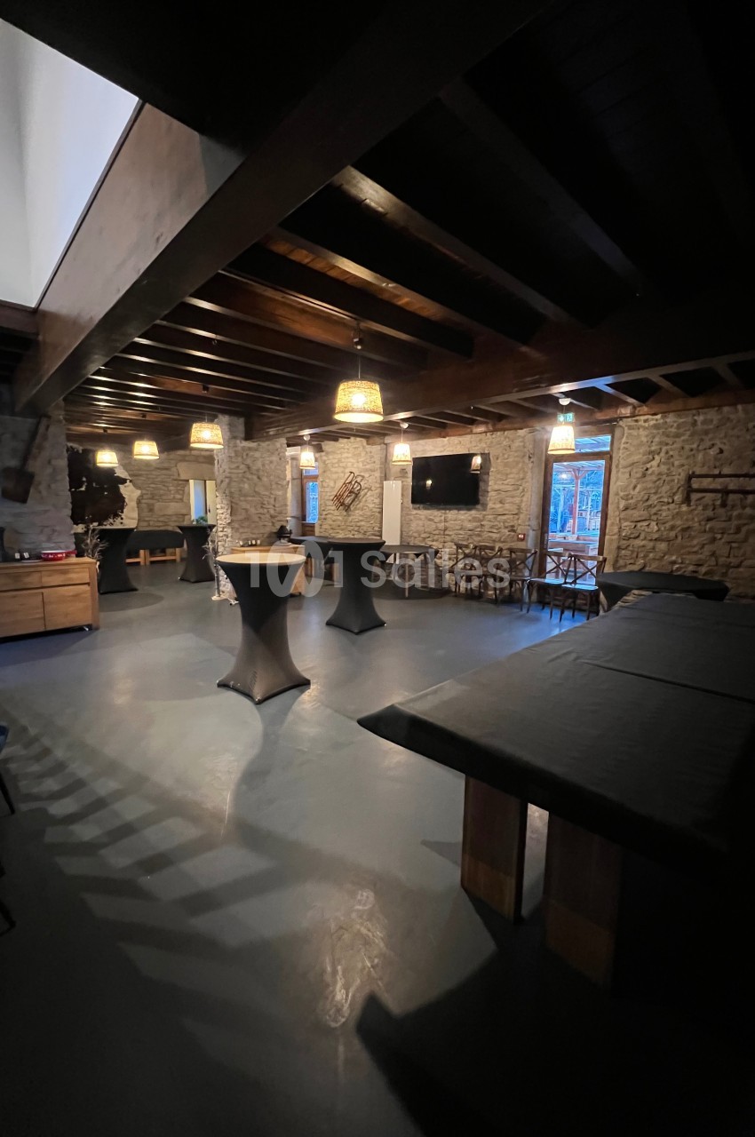 Salle intérieure avec murs en pierre, tables hautes, éclairage tamisé et grandes fenêtres donnant sur l'extérieur.