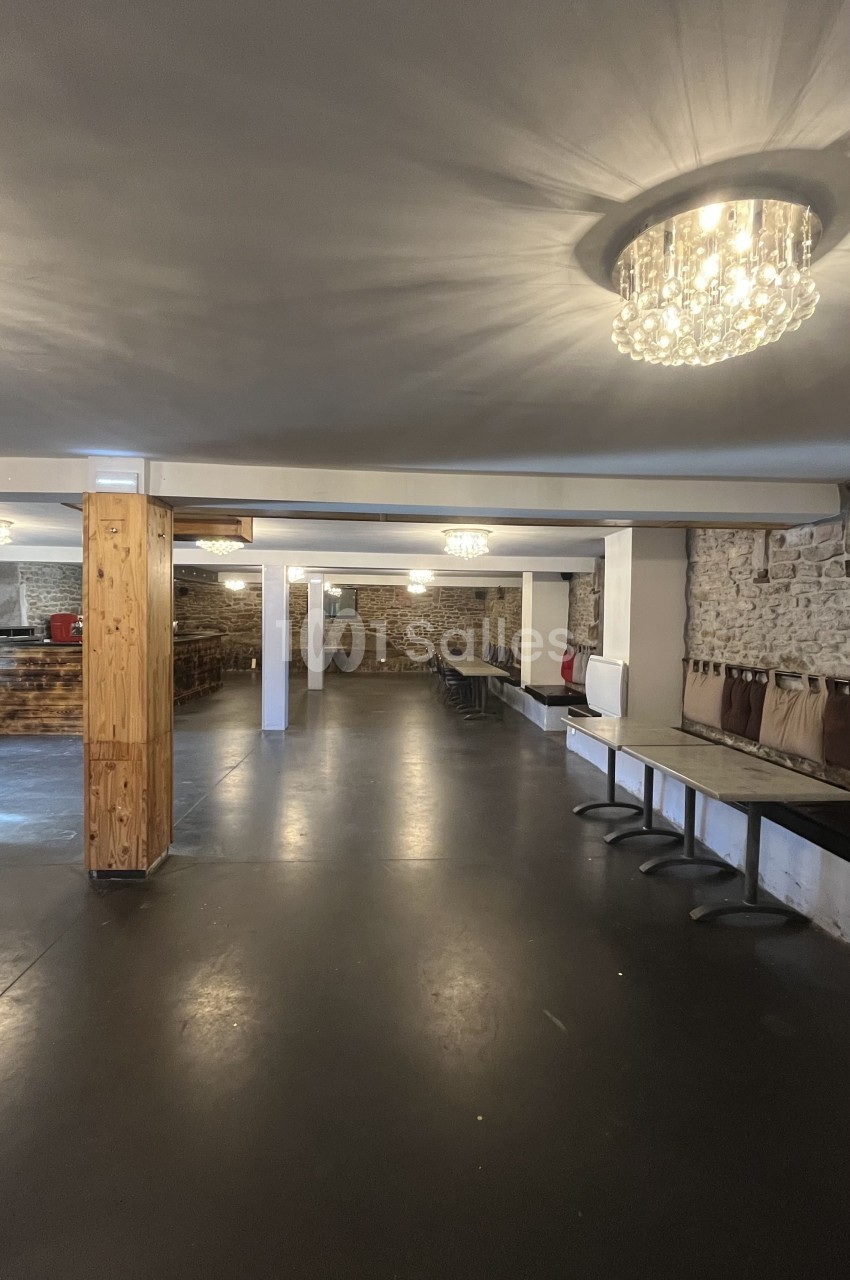 Salle vide avec sol en béton, murs en pierre et bois, tables pliées contre le mur et lustres suspendus au plafond.