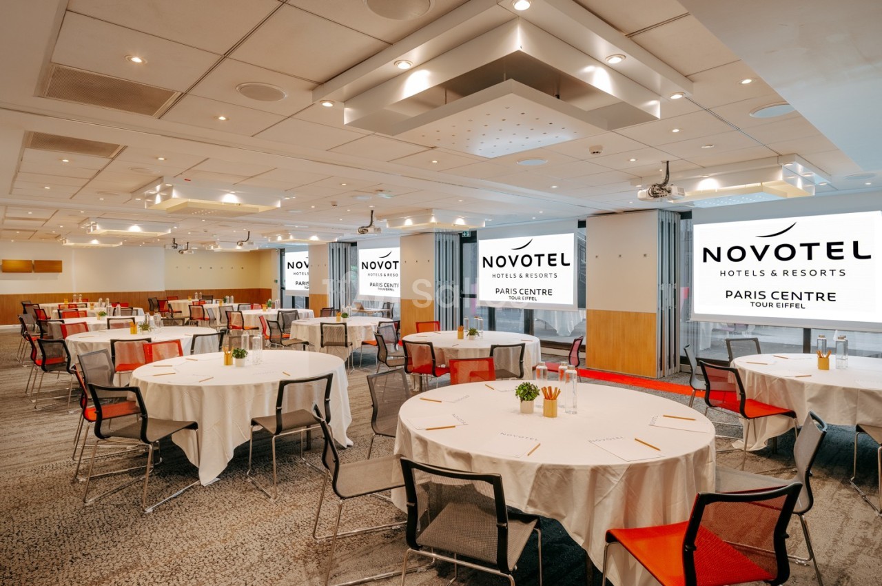 Salle de conférence lumineuse avec tables rondes dressées, chaises colorées et écrans affichant le logo Novotel.