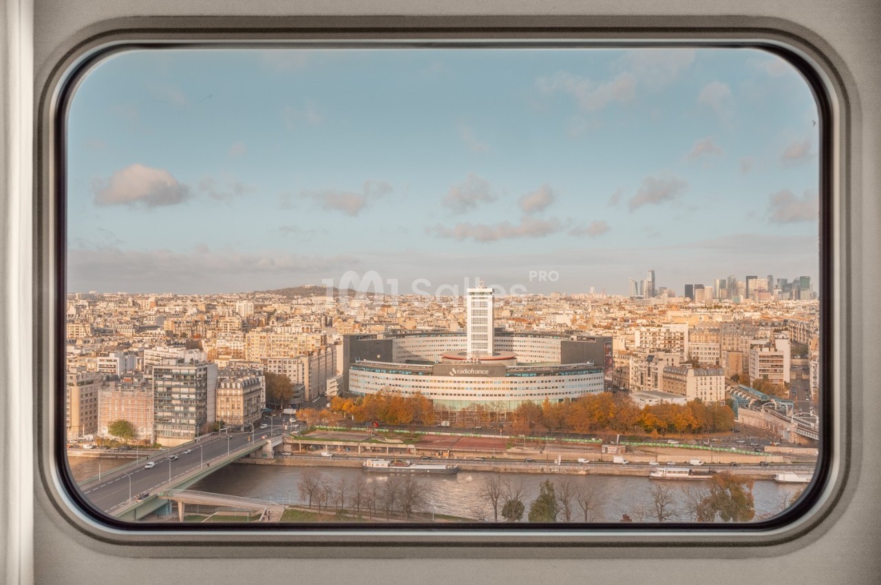 Vue de Paris à travers une fenêtre, montrant la Seine, un bâtiment moderne et l'horizon urbain avec des gratte-ciels.