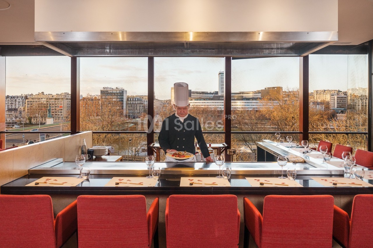 Un chef en tenue blanche présente un plat dans un restaurant avec vue sur une ville à travers de grandes baies vitrées.