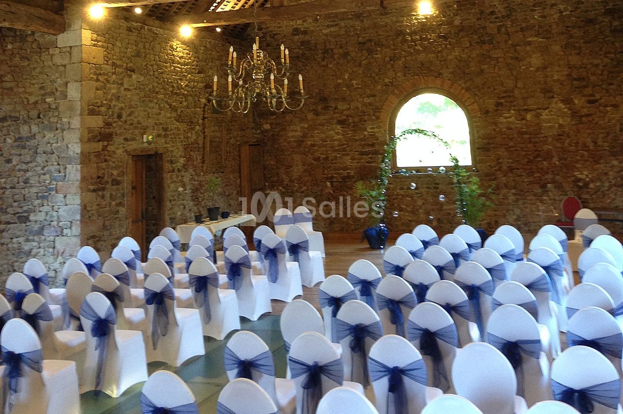 Salle en pierre avec des chaises blanches décorées de nœuds bleus, disposées pour un événement, sous un lustre suspendu.