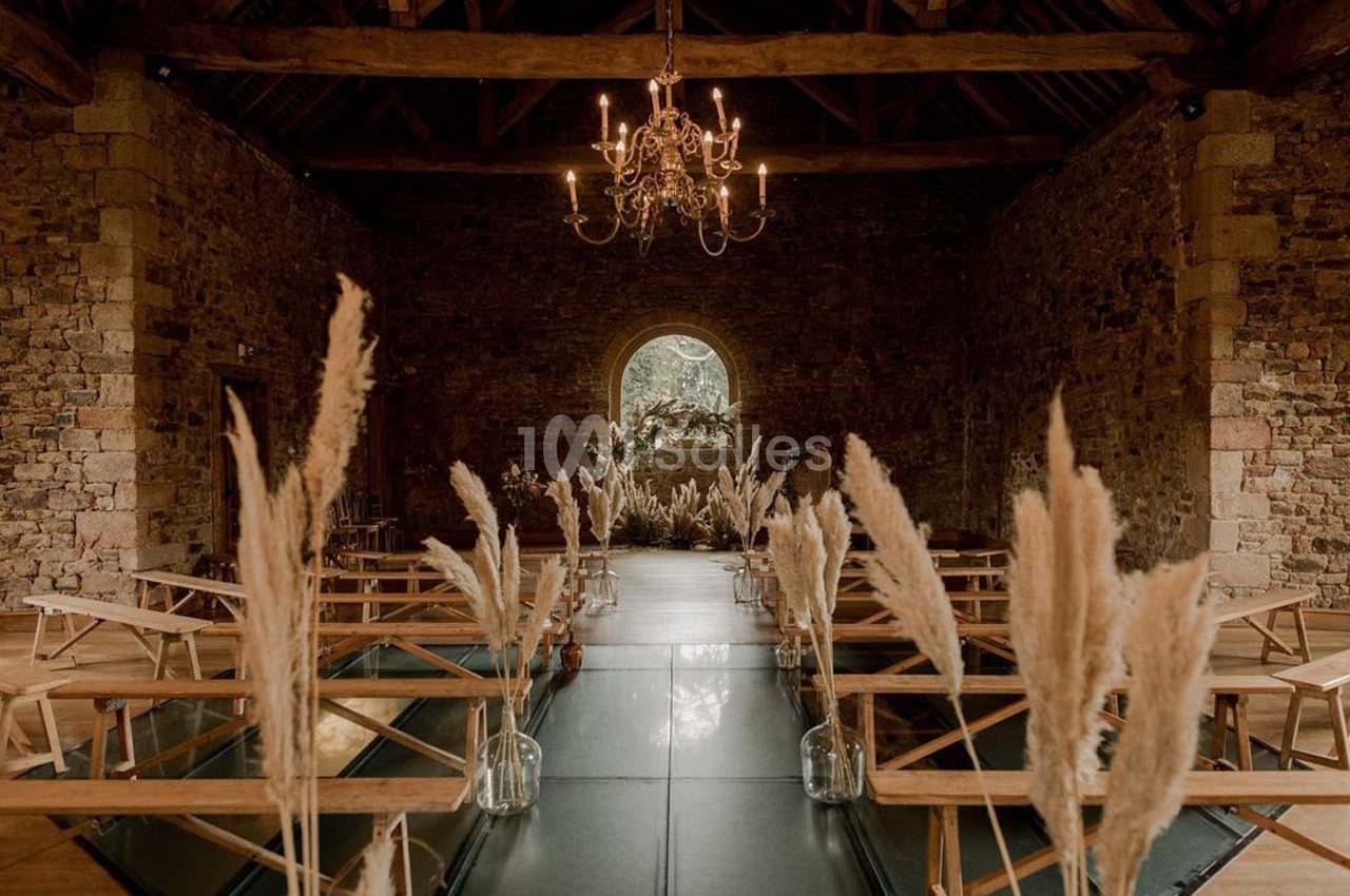 Salle en pierre avec poutres apparentes, décorée de pampas et bancs en bois, éclairée par un lustre central.