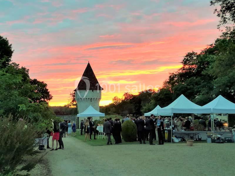 Groupe de personnes réunies près de tentes blanches dans un jardin au coucher du soleil, avec une tour en arrière-plan.