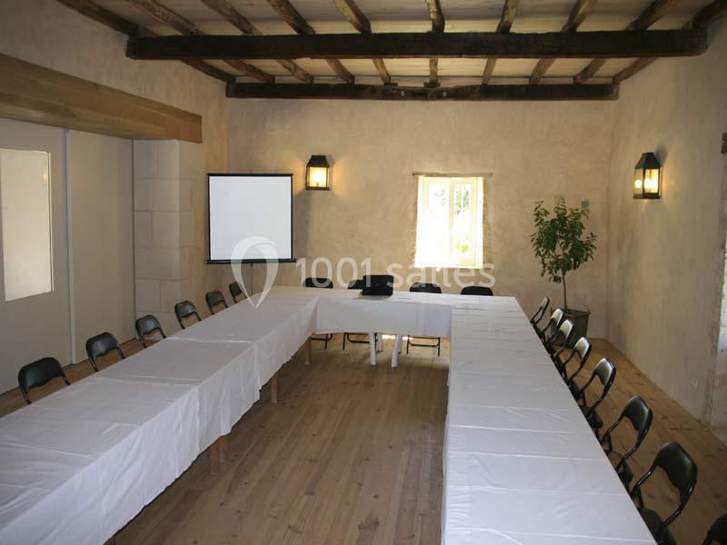 Salle de réunion lumineuse avec tables en U, chaises noires, projecteur et écran, murs clairs et poutres apparentes.
