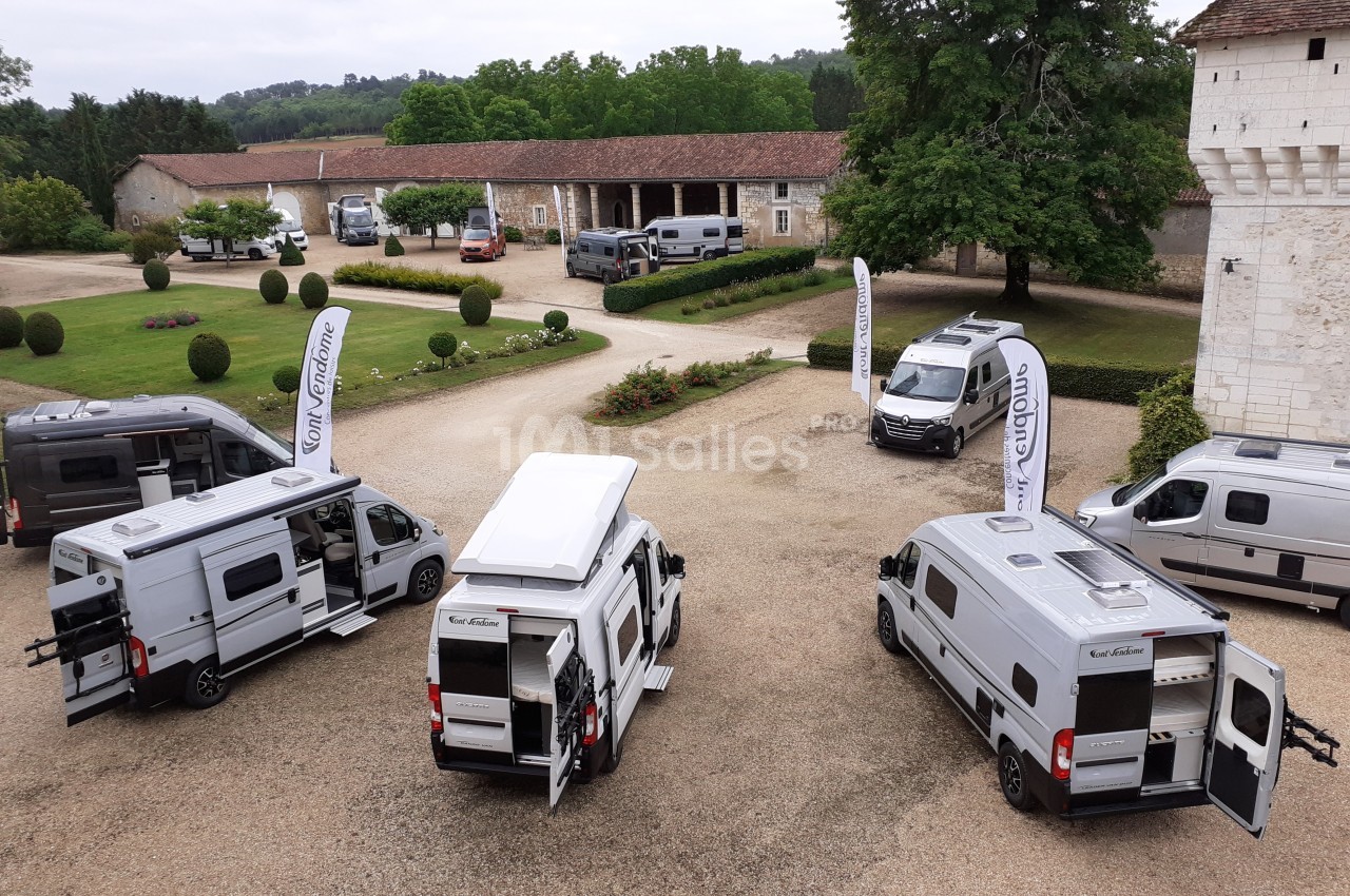 Plusieurs vans aménagés stationnés dans une cour pavée entourée de bâtiments et d'arbres.