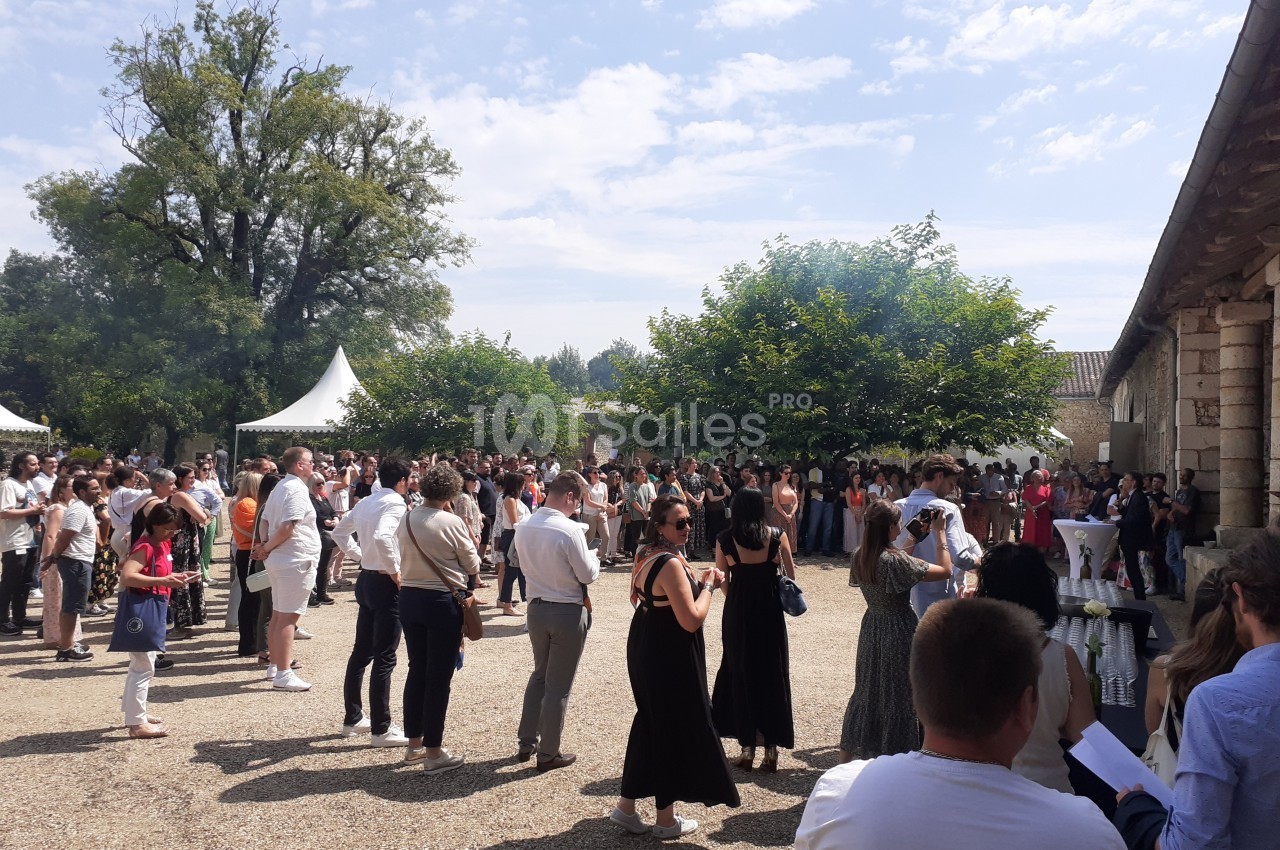Groupe de personnes rassemblées en extérieur dans une cour ensoleillée, entourée d'arbres et de tentes blanches.