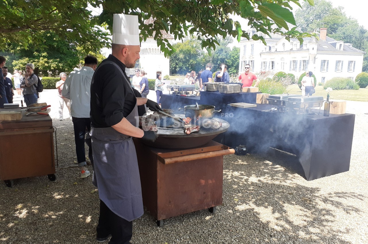 Un chef en tenue cuisine des plats sur une grande plancha en extérieur, entouré de convives et d'arbres.