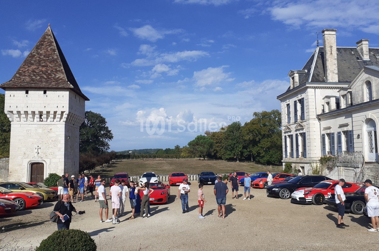 Rassemblement de voitures de sport devant un château et une tour, avec des visiteurs en plein air.