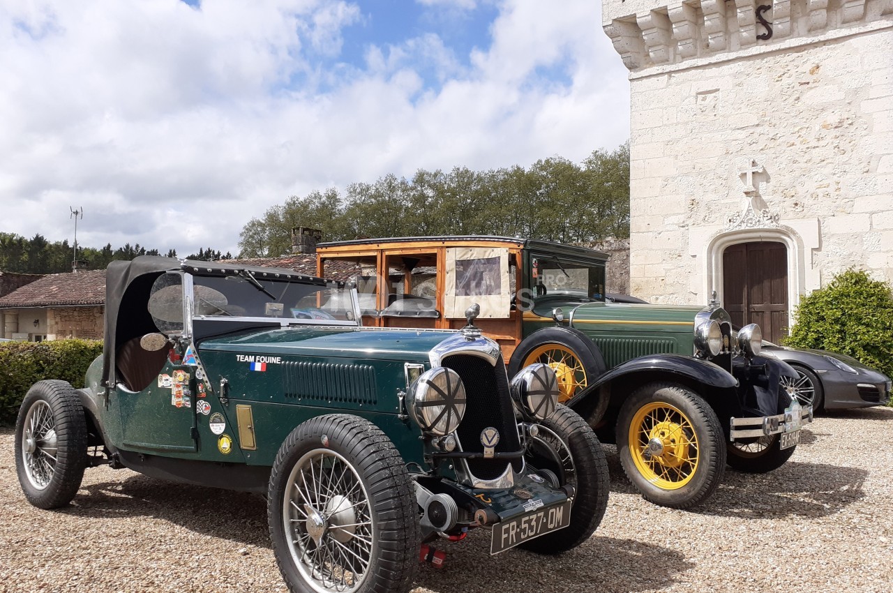 Deux voitures anciennes, une décapotable verte et une berline, stationnées devant un bâtiment en pierre.