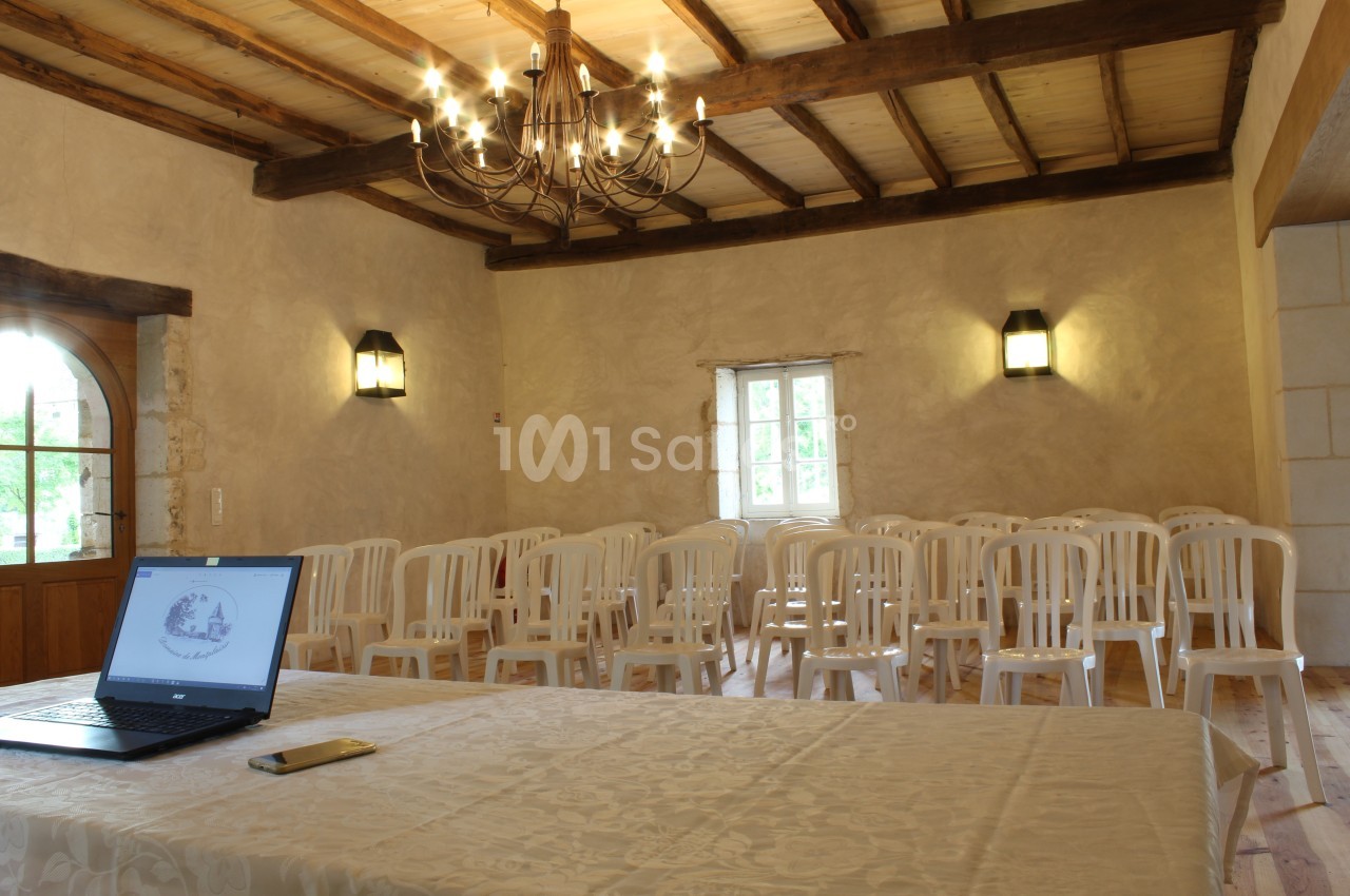 Salle lumineuse avec des chaises blanches alignées, une table recouverte d'une nappe et un ordinateur portable ouvert.