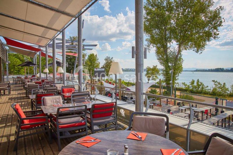 Terrasse ensoleillée avec tables dressées, chaises rouges et vue sur un lac entouré d'arbres.