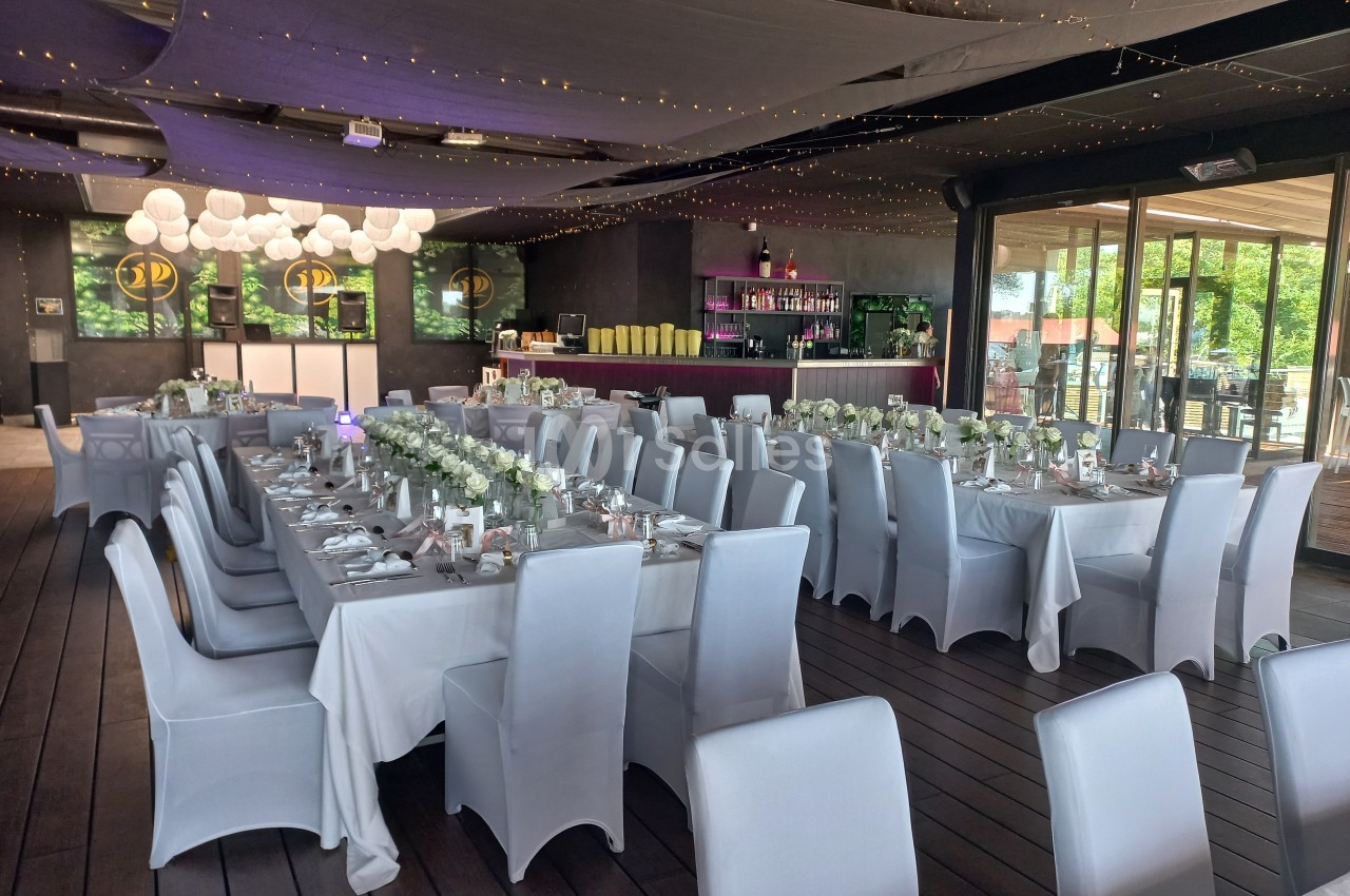 Salle de réception décorée avec des tables dressées, nappes blanches, chaises houssées et guirlandes lumineuses suspendues.