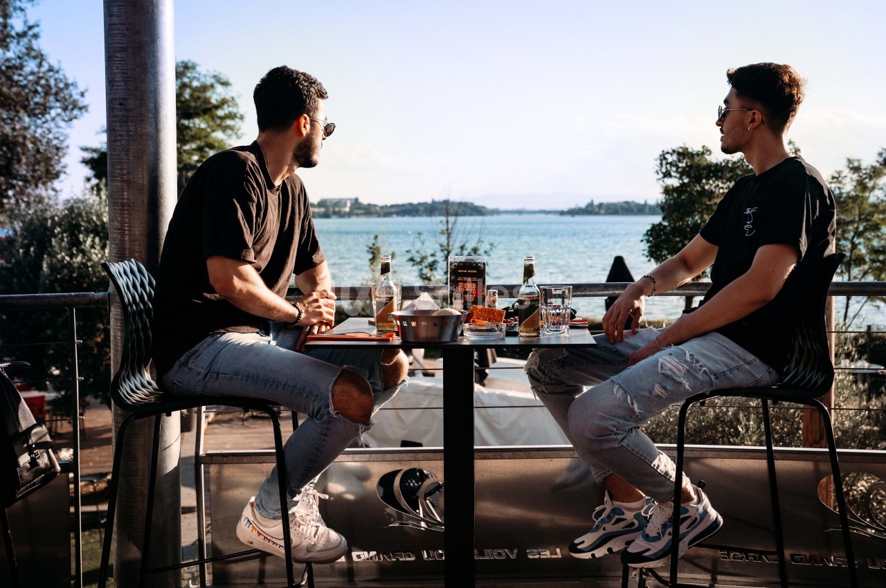 Deux hommes assis à une table en terrasse, partageant un repas avec vue sur un lac et des arbres en arrière-plan.