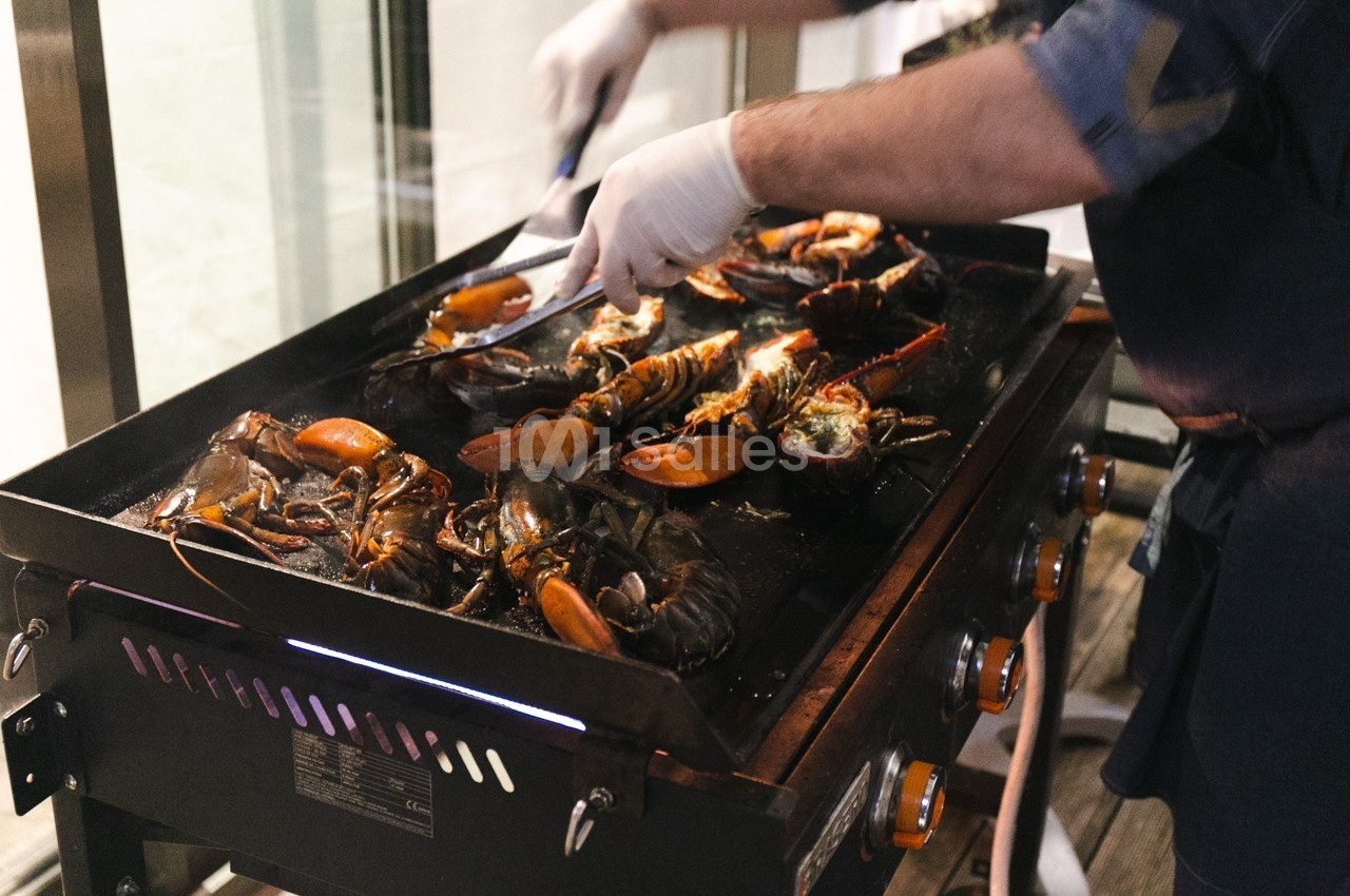 Personne en train de griller des homards sur une plancha, avec des pinces et des gants pour manipuler les crustacés.