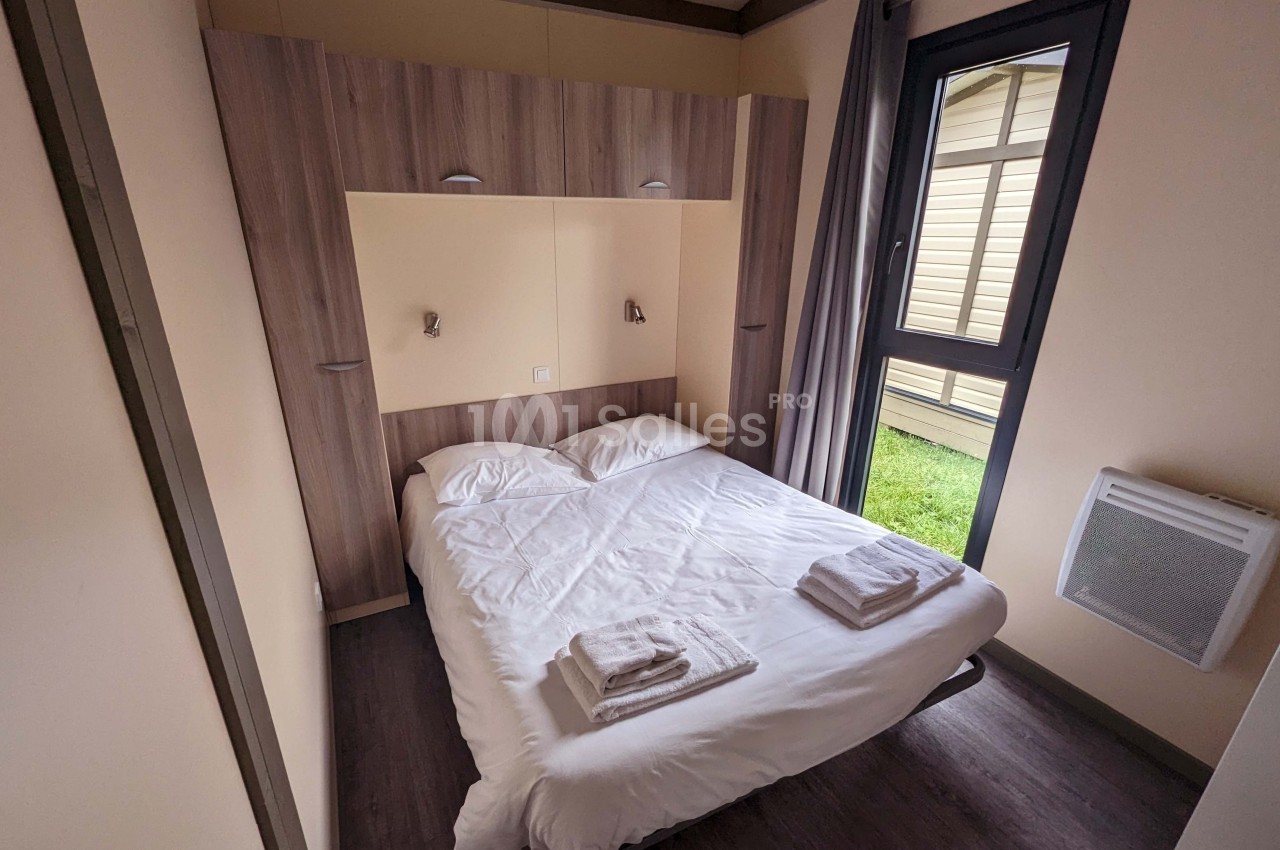 Chambre avec un lit double, draps blancs, serviettes pliées, mobilier en bois et fenêtre donnant sur un jardin.
