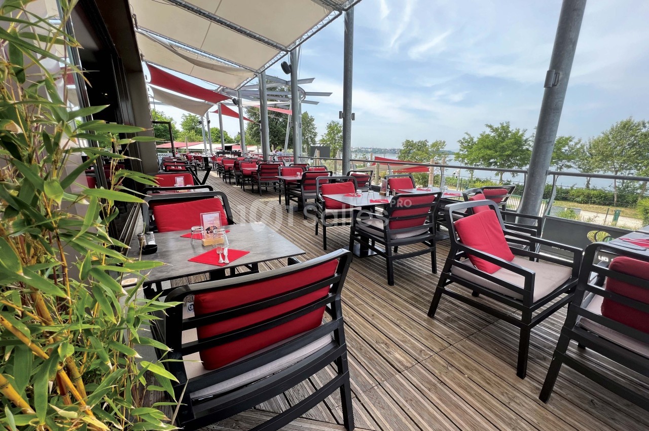 Terrasse d'un restaurant avec des tables et chaises rouges, vue sur un paysage arboré et un plan d'eau.