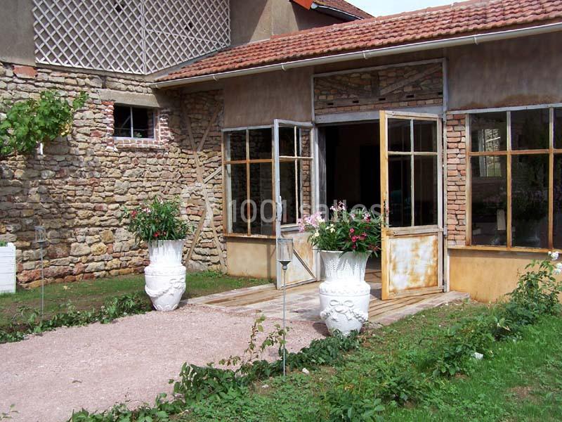 Entrée d'une maison en pierre avec des portes vitrées ouvertes, flanquée de deux grands pots de fleurs blanches.