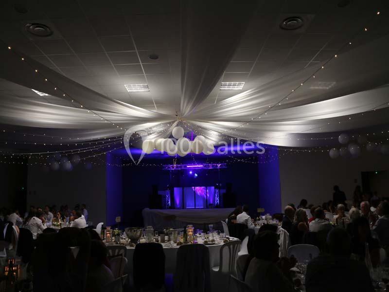 Salle de réception décorée avec des guirlandes lumineuses, des ballons suspendus et une scène éclairée en violet.