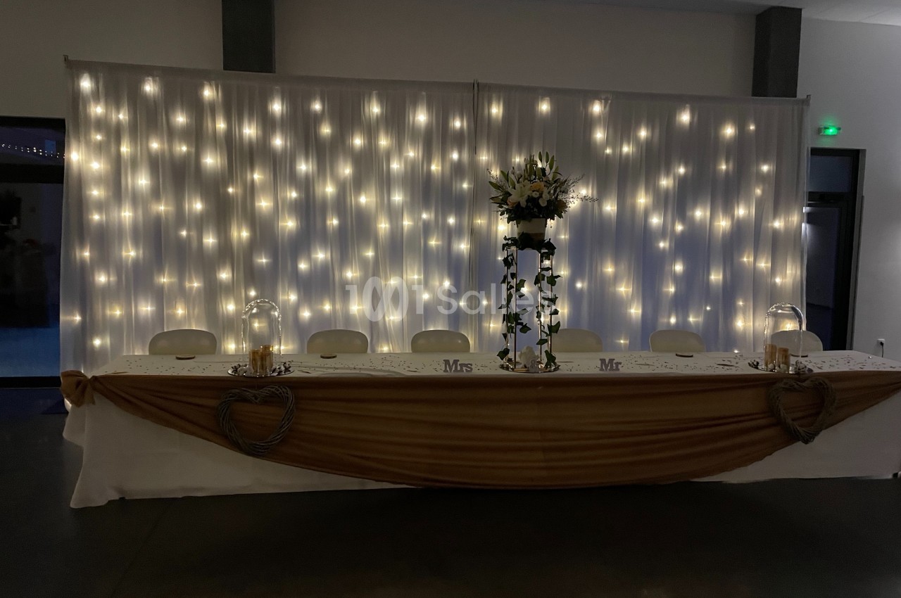 Table de mariage décorée avec une nappe dorée, des fleurs, des bougies et un fond lumineux de guirlandes LED.