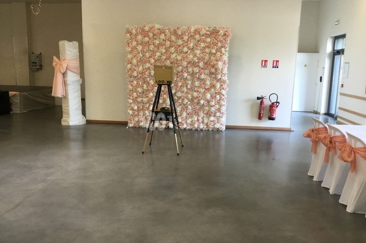 Mur de fleurs roses avec un photobooth sur trépied dans une salle décorée pour un événement.