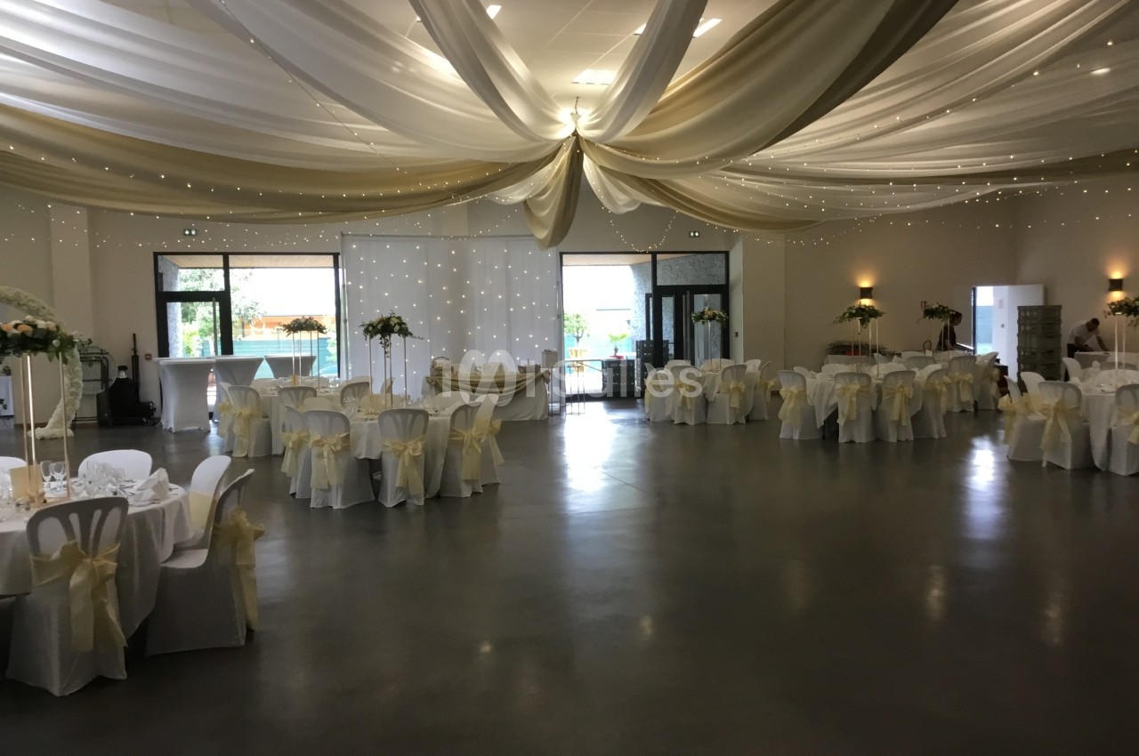 Salle de réception décorée avec des tables rondes, nappes blanches, chaises ornées de nœuds et drapés au plafond.