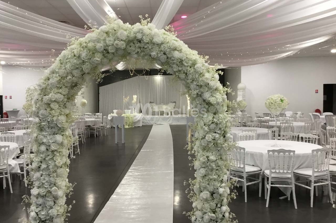 Salle de réception décorée pour un mariage avec une arche de fleurs blanches, tables rondes et chaises blanches.