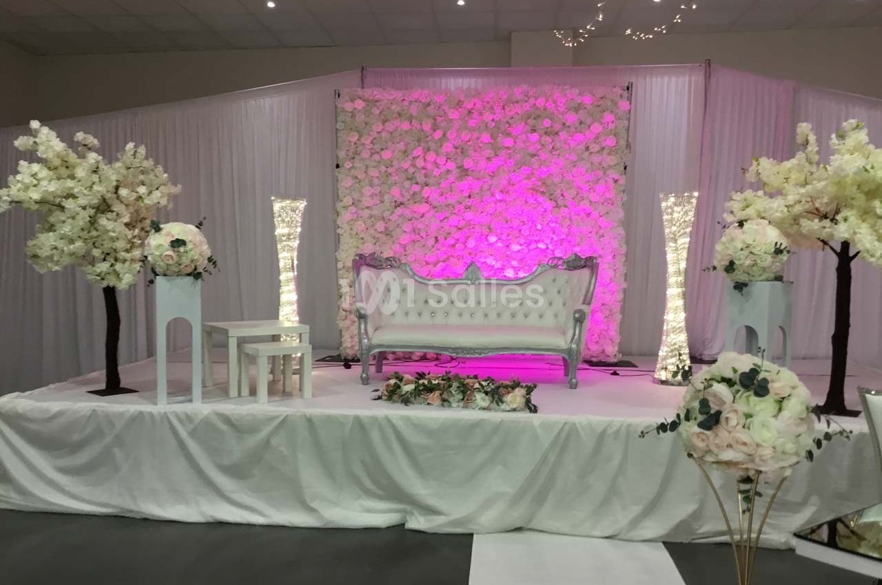 Décor de scène avec canapé blanc, murs fleuris, colonnes lumineuses et arrangements floraux dans une ambiance rose.