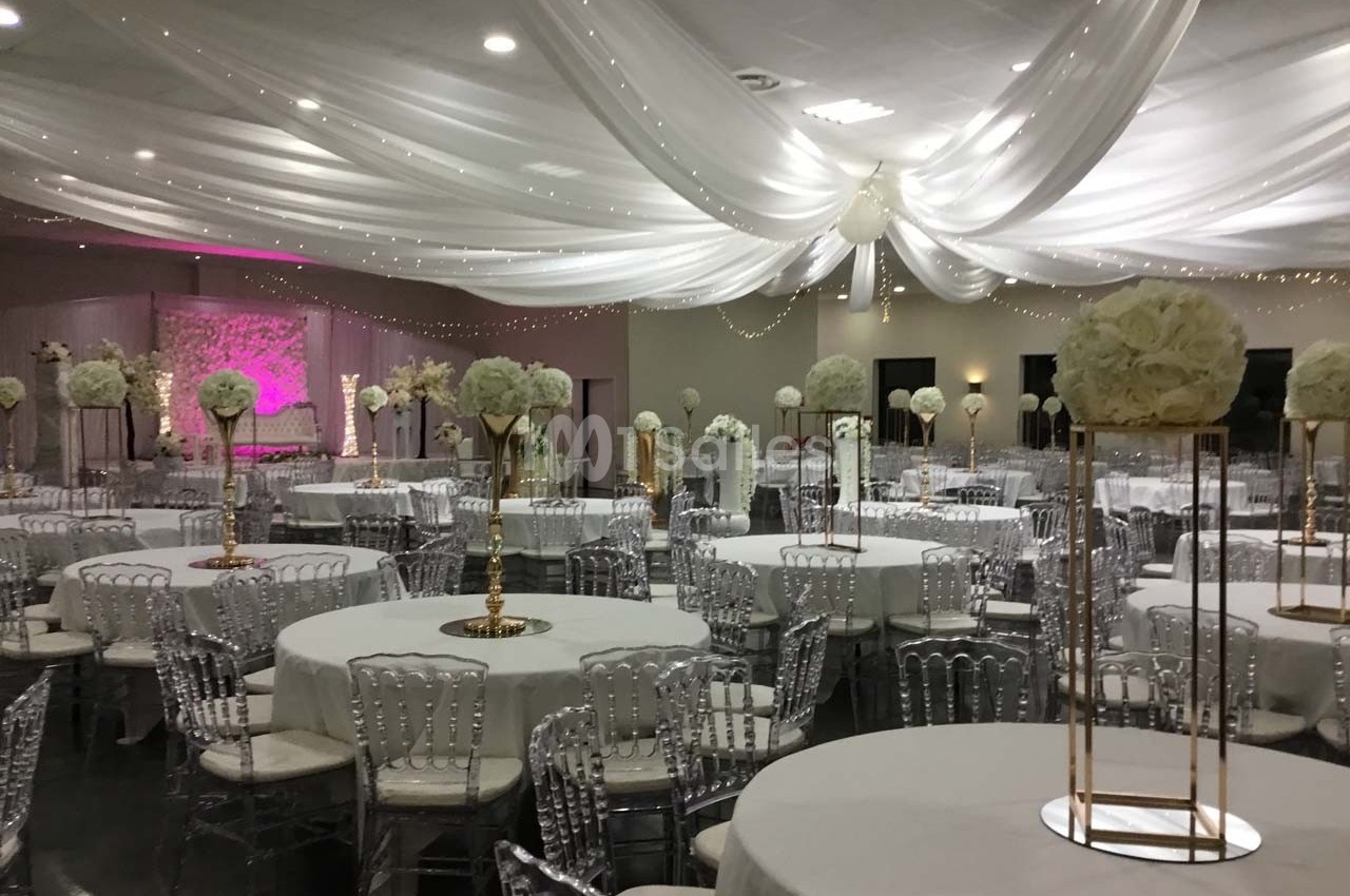 Salle de réception décorée avec des tables rondes, nappes blanches, chaises transparentes et centres de table fleuris.