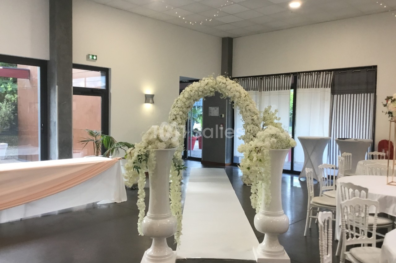 Arche de fleurs blanches décorative au centre d'une salle de réception lumineuse avec tables et chaises blanches.