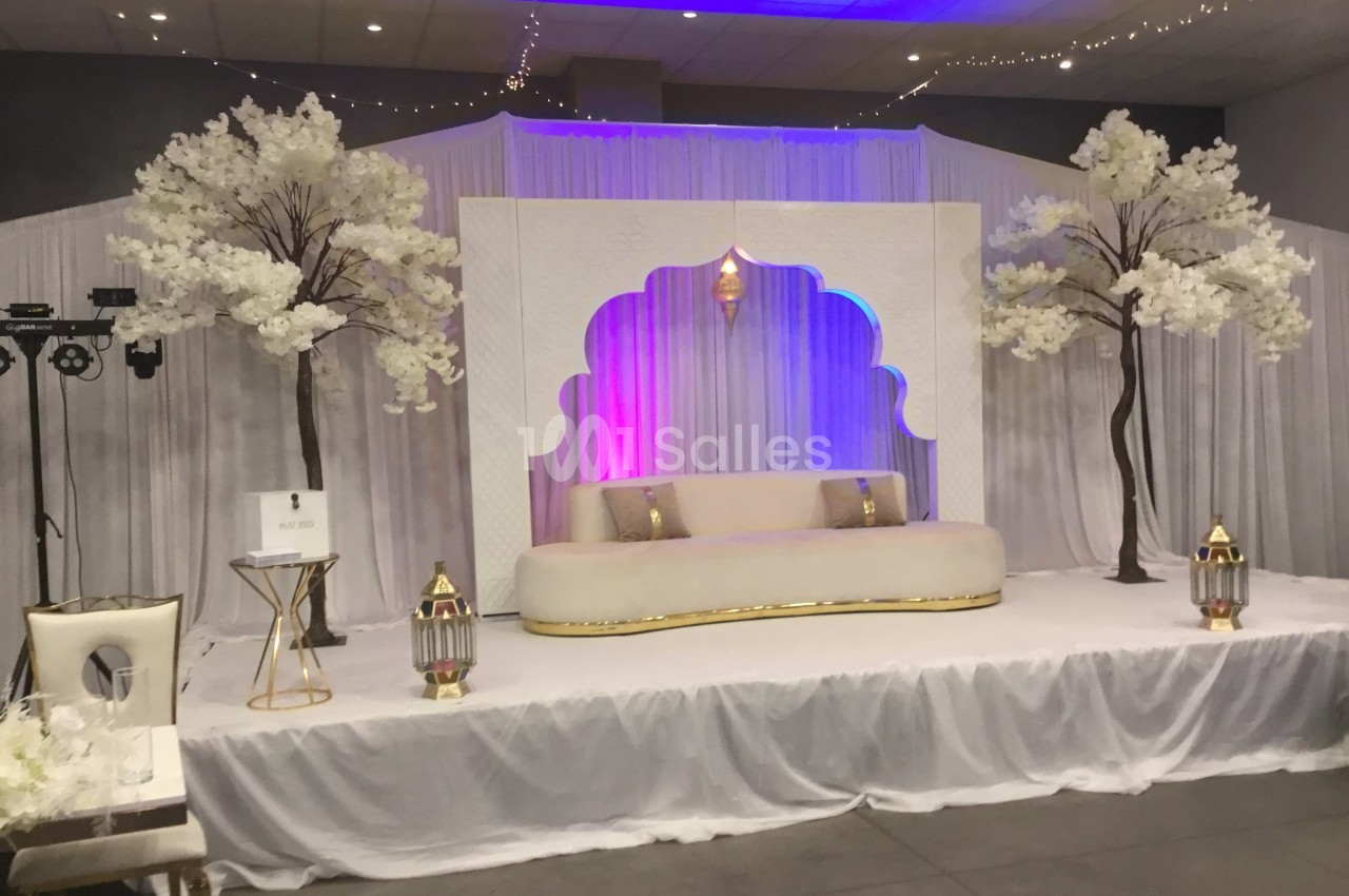 Décor de scène avec canapé beige, lanternes dorées, rideaux blancs et arbres artificiels ornés de fleurs blanches.